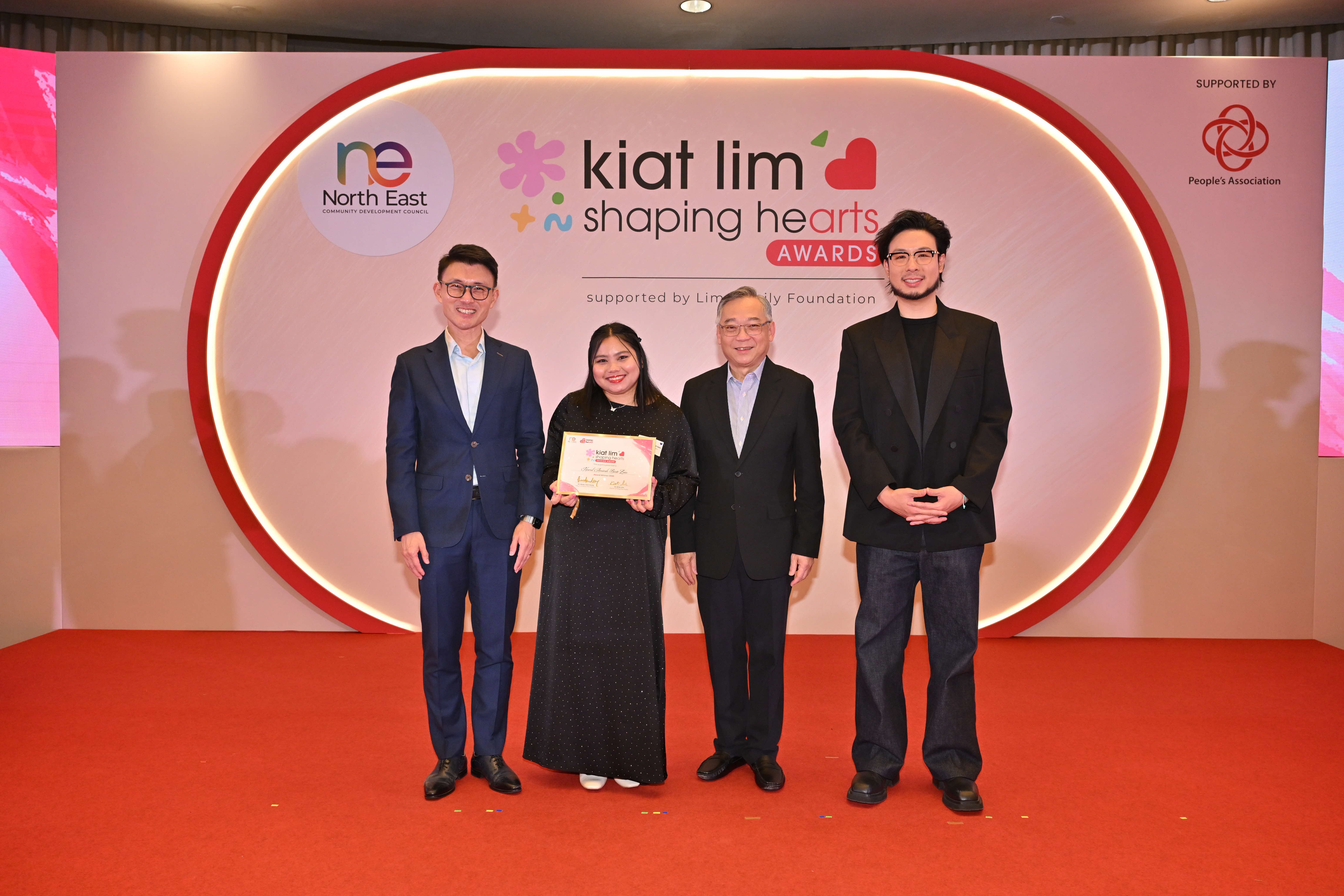 Award recipient standing on stage with the presenter, holding their certificate and posing for a group photo during the award ceremony, with a backdrop reading ‘Kiat Lim-Shaping Hearts Awards'.